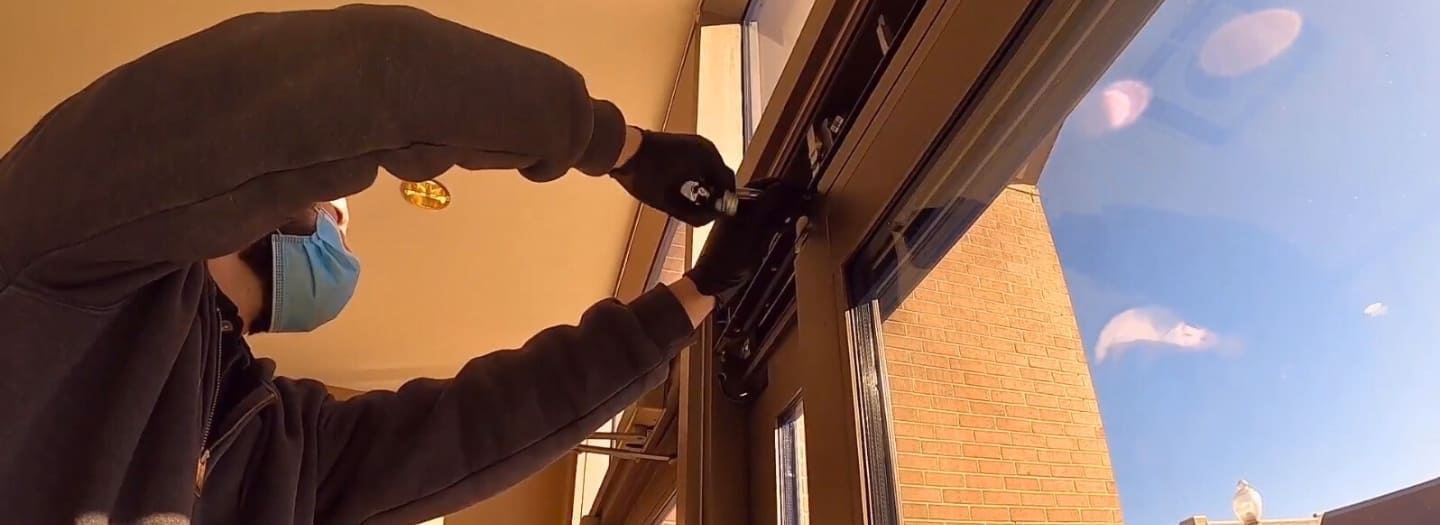 A person wearing a mask and gloves adjusts the mechanism of a large window from inside the building, ensuring seamless operation alongside routine commercial door repair tasks.