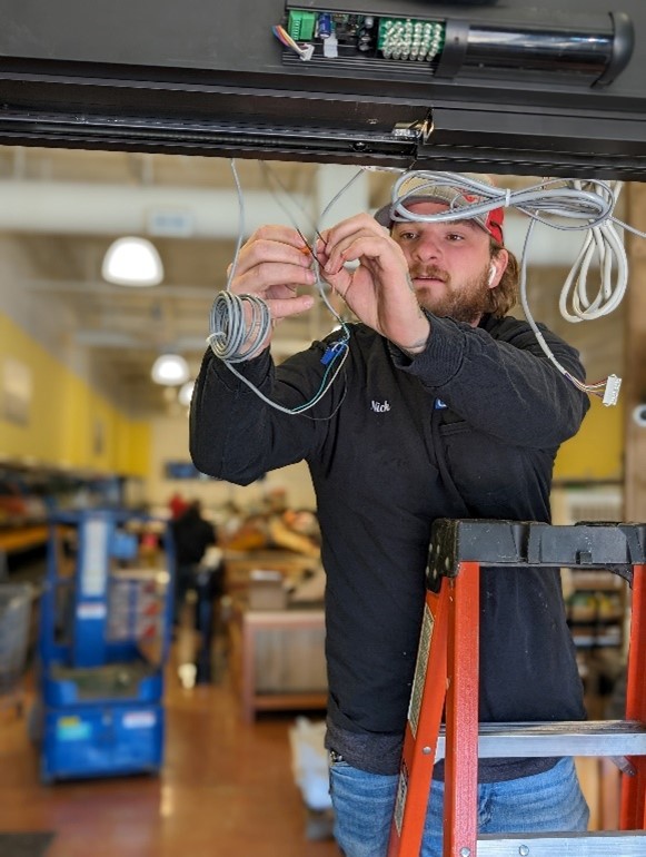 Person on a ladder installing cables in a store, wearing a black jacket and red cap, as part of an automatic door service to enhance accessibility and convenience for customers.